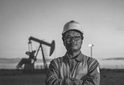 An oil and gas worker wearing a safety helmet and uniform stands in front of an oil pump jack at dusk.